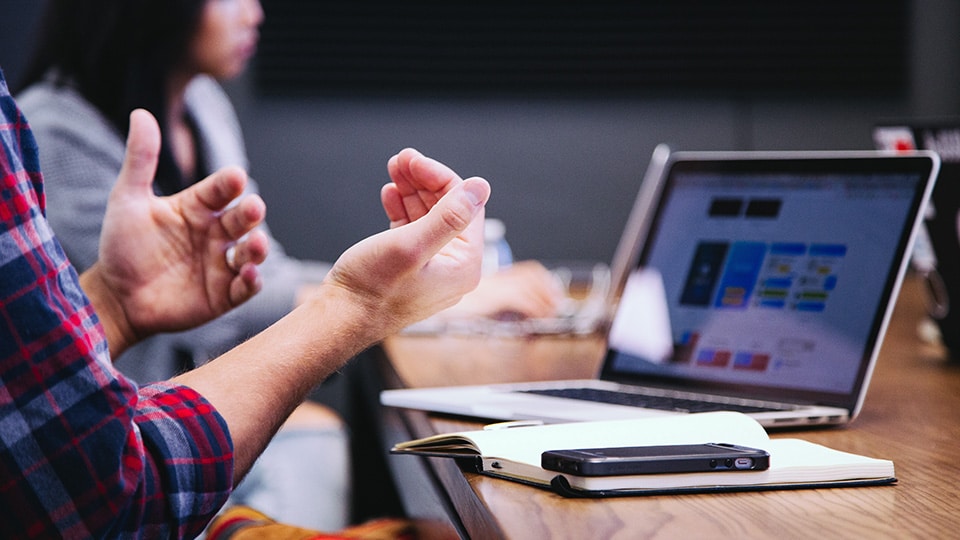 Photo of person explaining something to another in front of a open laptop on a desk, with a book and phone as a paper weight.